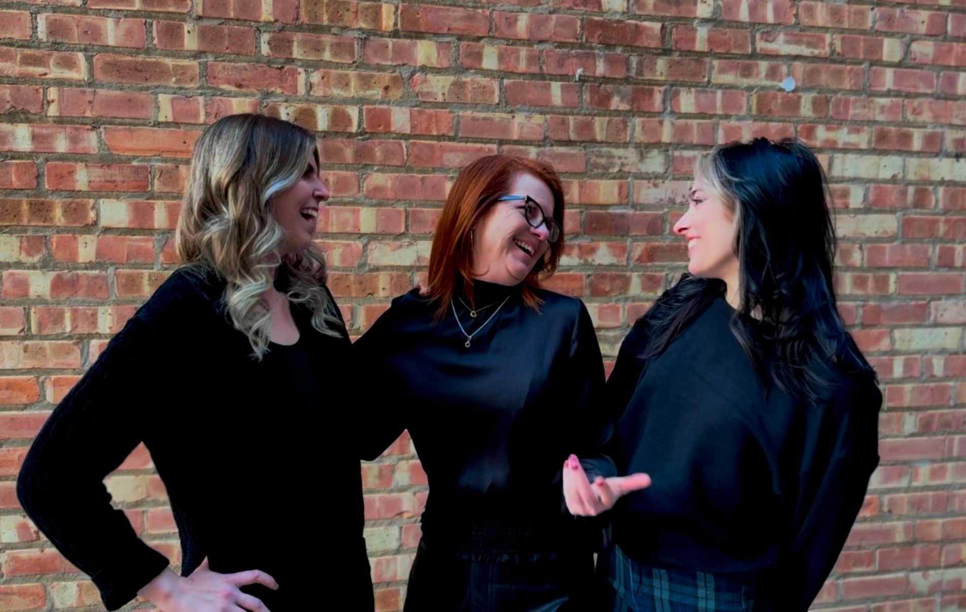 Three women in black tops laughing and chatting in front of a brick wall.
